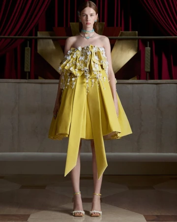 Model posing in a structured yellow mini dress with floral applique and high jewelry in front of red theatre curtains.