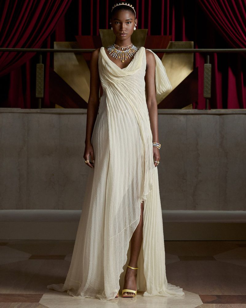Model wearing a draped cream pleated gown with an ornate high jewelry necklace and gold heels in front of red velvet curtains.