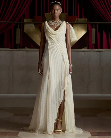Model wearing a draped cream pleated gown with an ornate high jewelry necklace and gold heels in front of red velvet curtains.