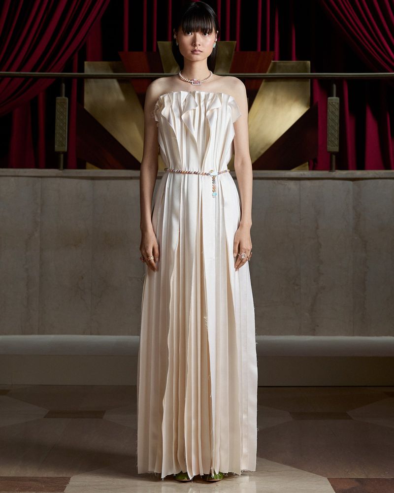 East Asian model in a strapless ivory pleated gown and delicate jewelry standing before draped red velvet curtains.