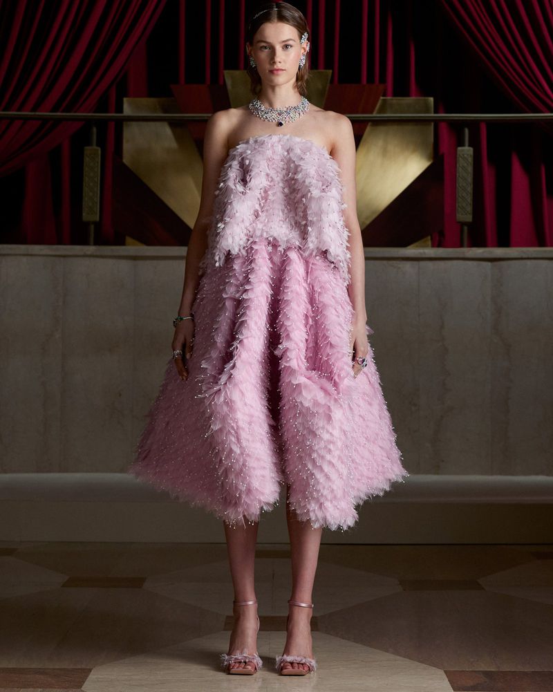 Model wearing an ornate diamond necklace and a strapless pink feathered midi dress against deep red curtains and a gold backdrop.
