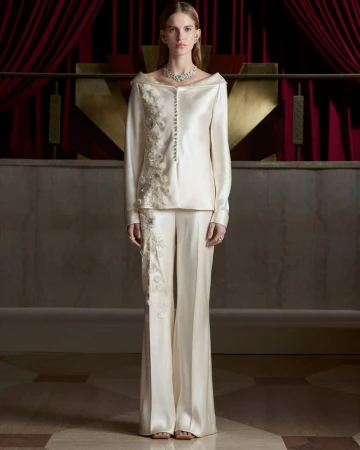 Model wearing an ivory satin embroidered pantsuit and high jewelry necklace in front of red draped curtains.