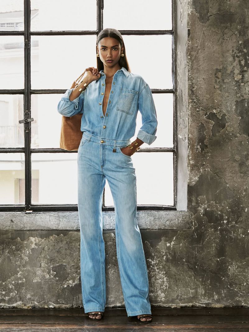 Model Radija T poses in a light wash denim jumpsuit holding a brown suede bag beside an industrial grid window.