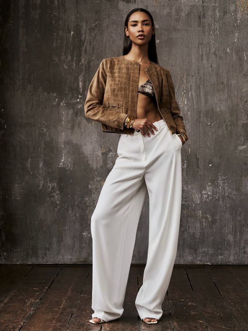 Model Radija T wearing a brown cropped jacket and wide-leg white trousers against a textured studio backdrop.