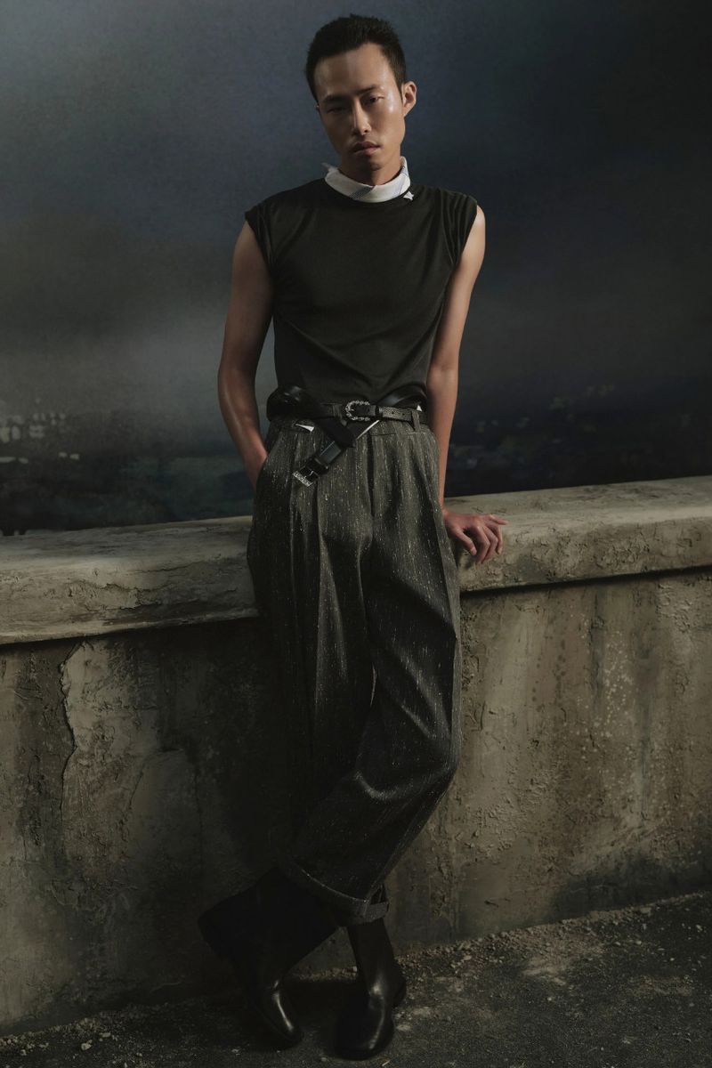 Model Jiseok Nam in a black sleeveless top and grey pleated trousers leans against a concrete ledge.