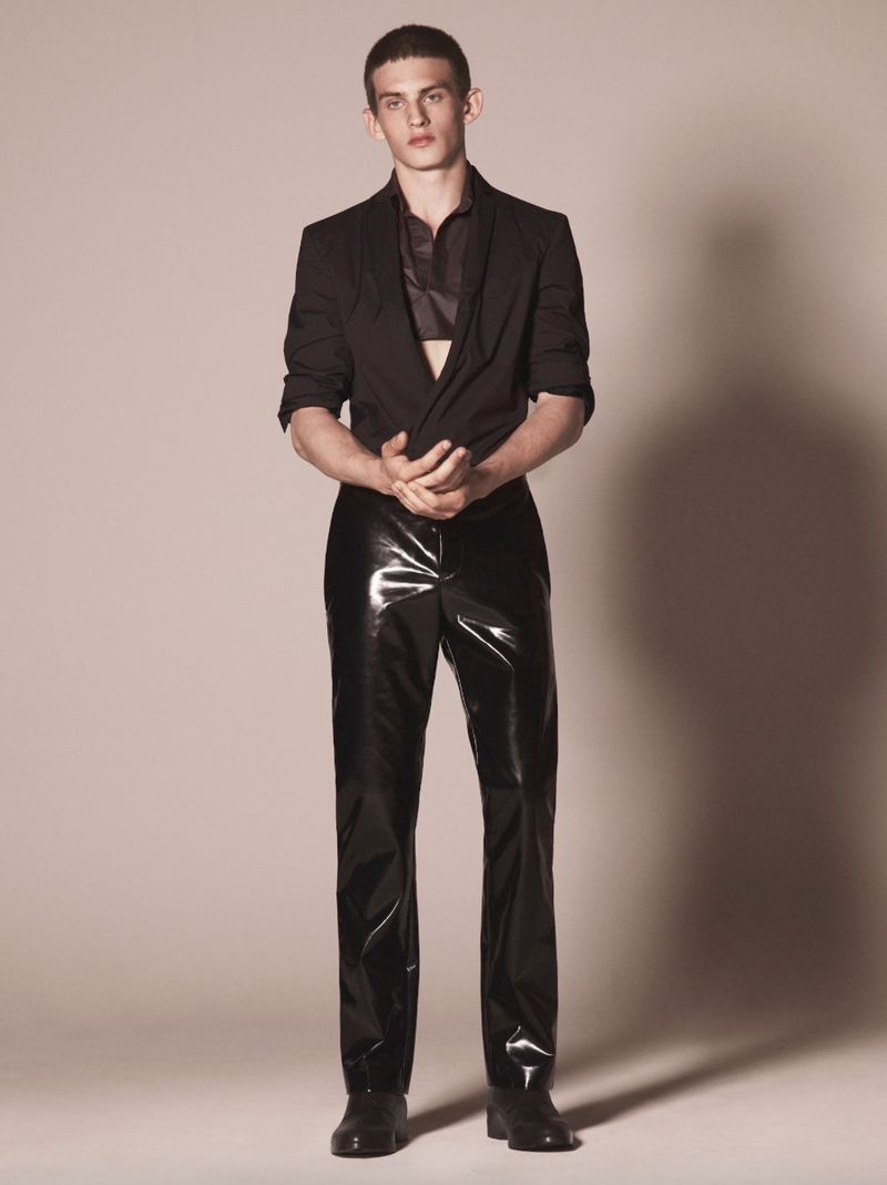 Model Joe Bottomer stands in a black jacket and glossy black vinyl trousers against a neutral studio backdrop.