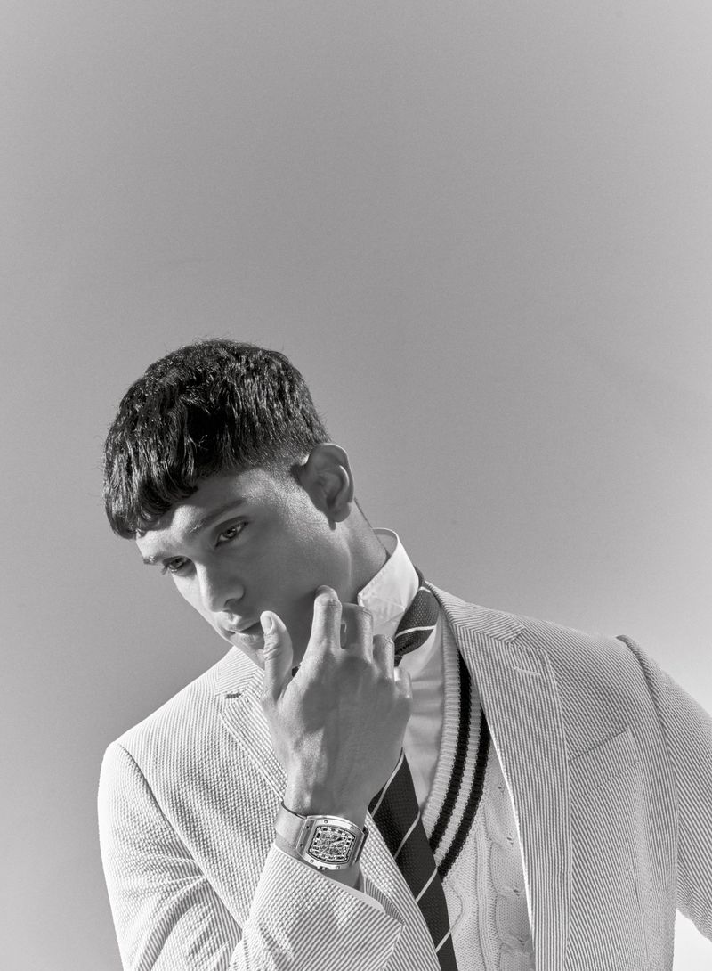 Monochrome portrait of a male model wearing a blazer and vest, hand near his face, with a wristwatch, against a light grey backdrop.