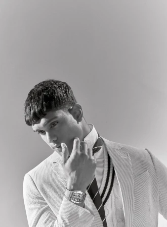 Monochrome portrait of a male model wearing a blazer and vest, hand near his face, with a wristwatch, against a light grey backdrop.