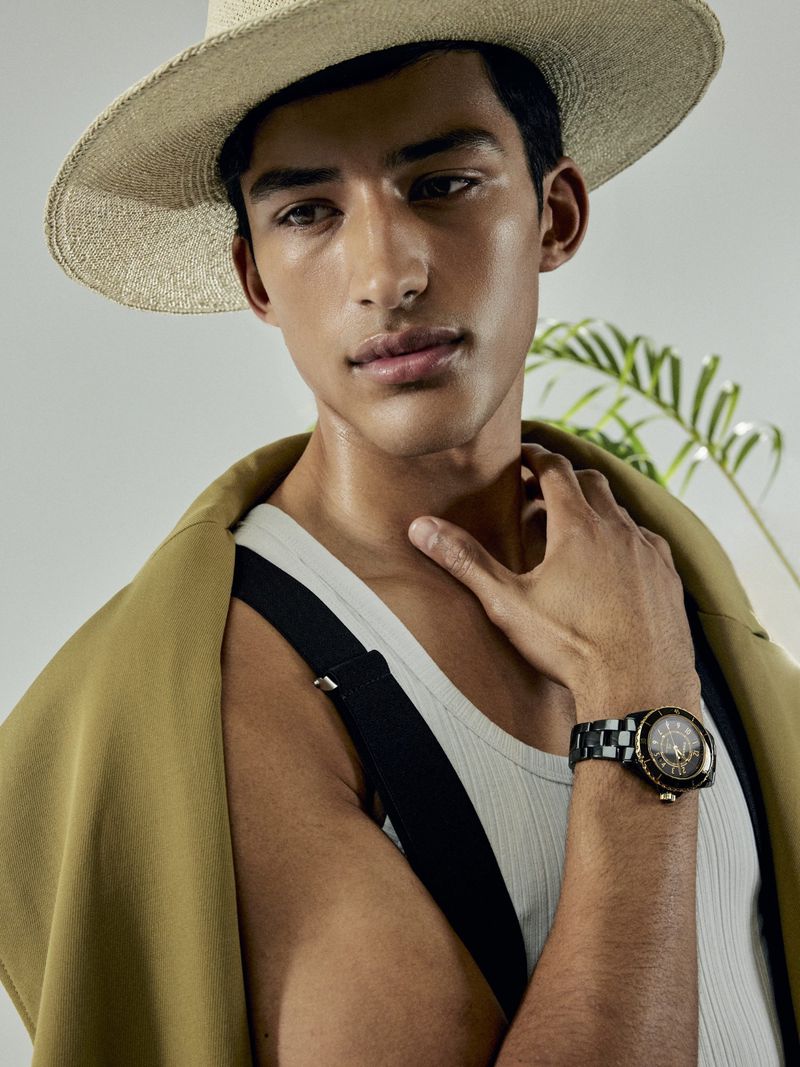 Portrait of a handsome man wearing a straw hat and wristwatch, posed against a neutral backdrop with a leafy plant in the background.