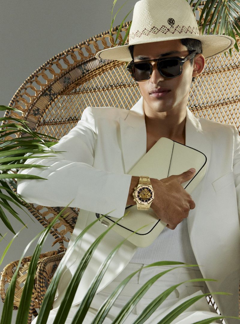 A man poses in a woven chair, wearing a white suit, straw hat, and sunglasses, holding a clutch bag, set against a grey background.