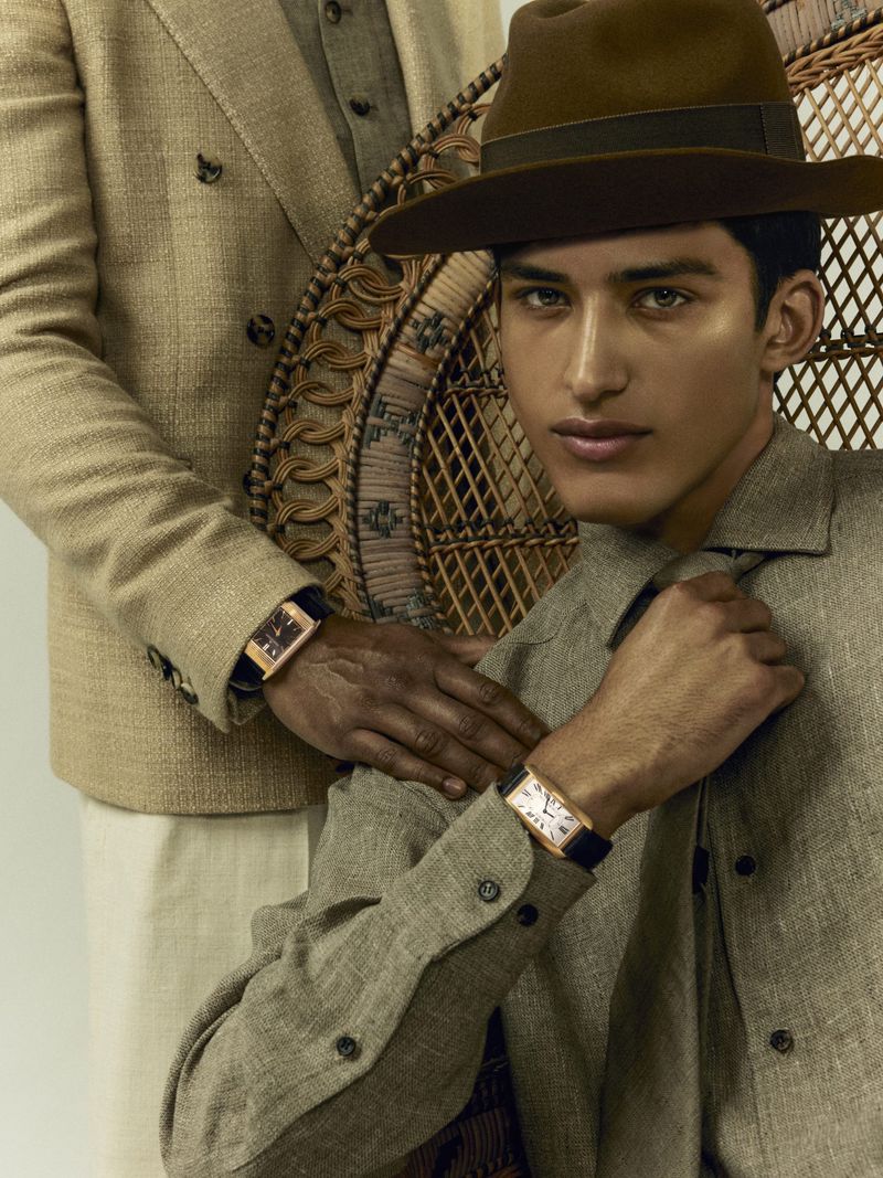 Two men pose in coordinated linen outfits and watches. One sits in a wicker chair, the other stands and places a hand on the seated man's shoulder.