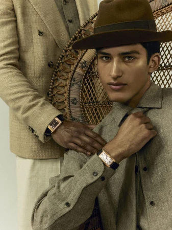 Two men pose in coordinated linen outfits and watches. One sits in a wicker chair, the other stands and places a hand on the seated man's shoulder.