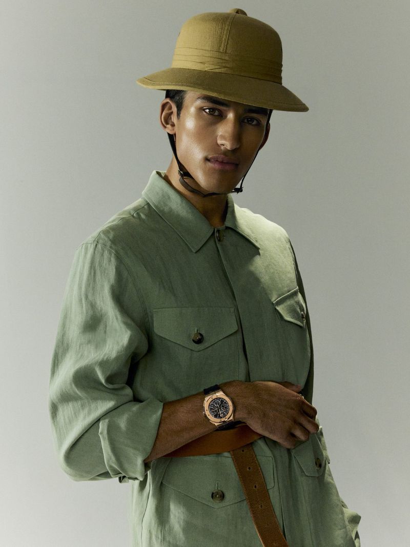 Portrait of a man in safari hat and green utility suit with a watch on wrist, posing in front of a neutral gray backdrop.