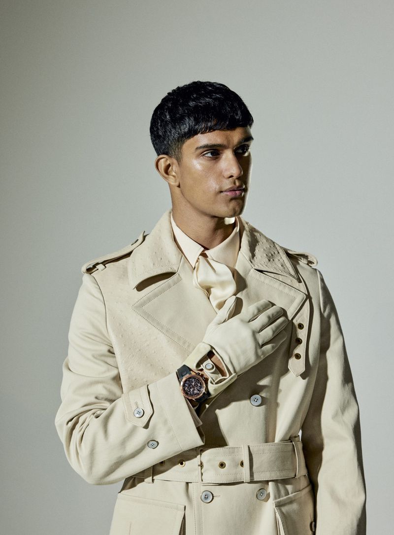 A man in a beige trench coat poses in a studio setting, wearing a watch and gloves. Fashion editorial with soft lighting.