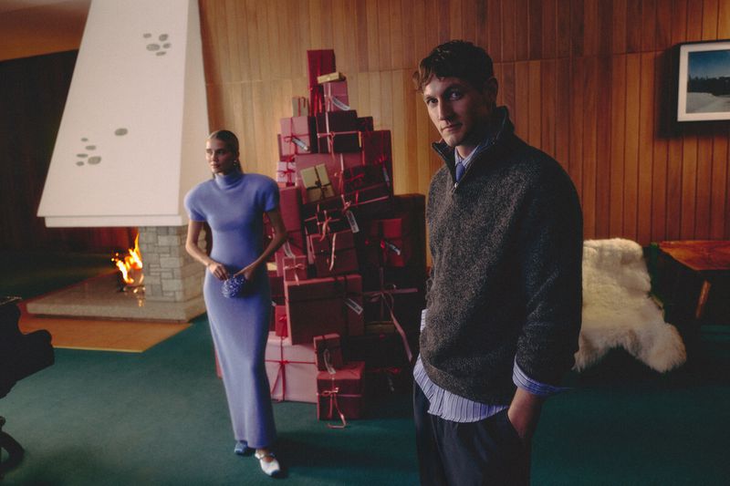 Rutger Schoone and a woman pose in a mid-century interior beside a tall stack of red Christmas gift boxes and a lit fireplace.