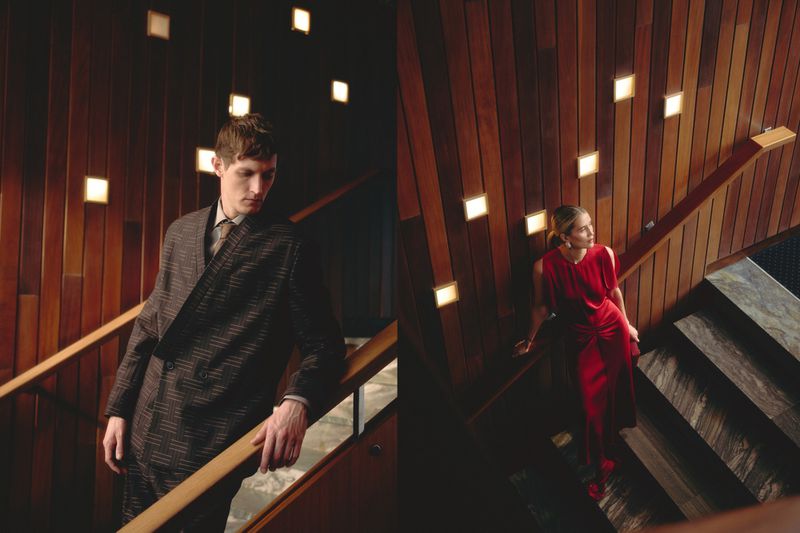 Rutger Schoone in a dark patterned suit alongside a woman in a red evening dress standing on a wood-paneled staircase with wall lights.
