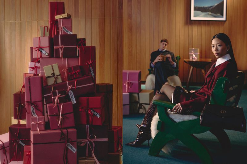 Asian woman in a burgundy blazer and boots sits next to a towering stack of pink and red gift boxes, with a man seated in the background.