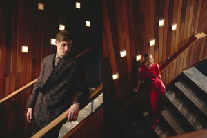 Rutger Schoone in a dark patterned suit alongside a woman in a red evening dress standing on a wood-paneled staircase with wall lights.