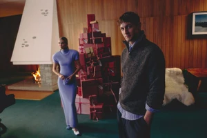 Rutger Schoone and a woman pose in a mid-century interior beside a tall stack of red Christmas gift boxes and a lit fireplace.
