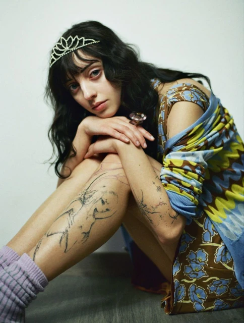 Young woman with tattoos, dark hair, and a tiara sits indoors, wearing a patterned top and lilac socks, against a light backdrop.