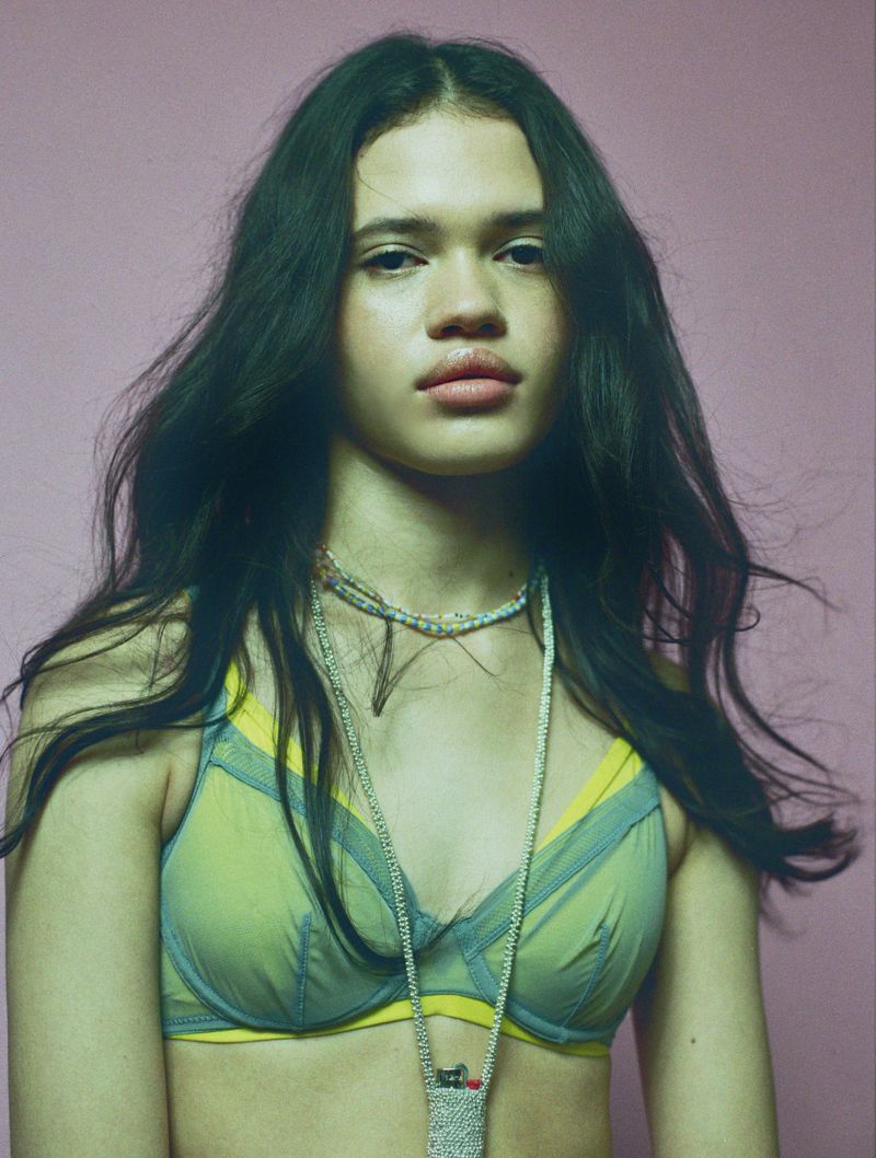 A young woman with long dark hair gazes directly at the viewer in a studio shot with a soft pink background, wearing a sheer green bra.