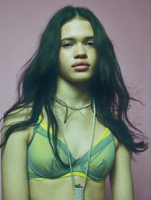 A young woman with long dark hair gazes directly at the viewer in a studio shot with a soft pink background, wearing a sheer green bra.