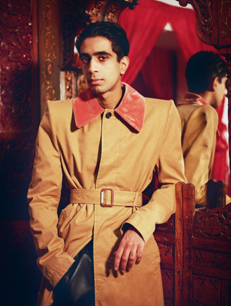 Stylish portrait of a young adult male in a beige trench coat with a pink collar, standing indoors with dark hair and brown eyes.
