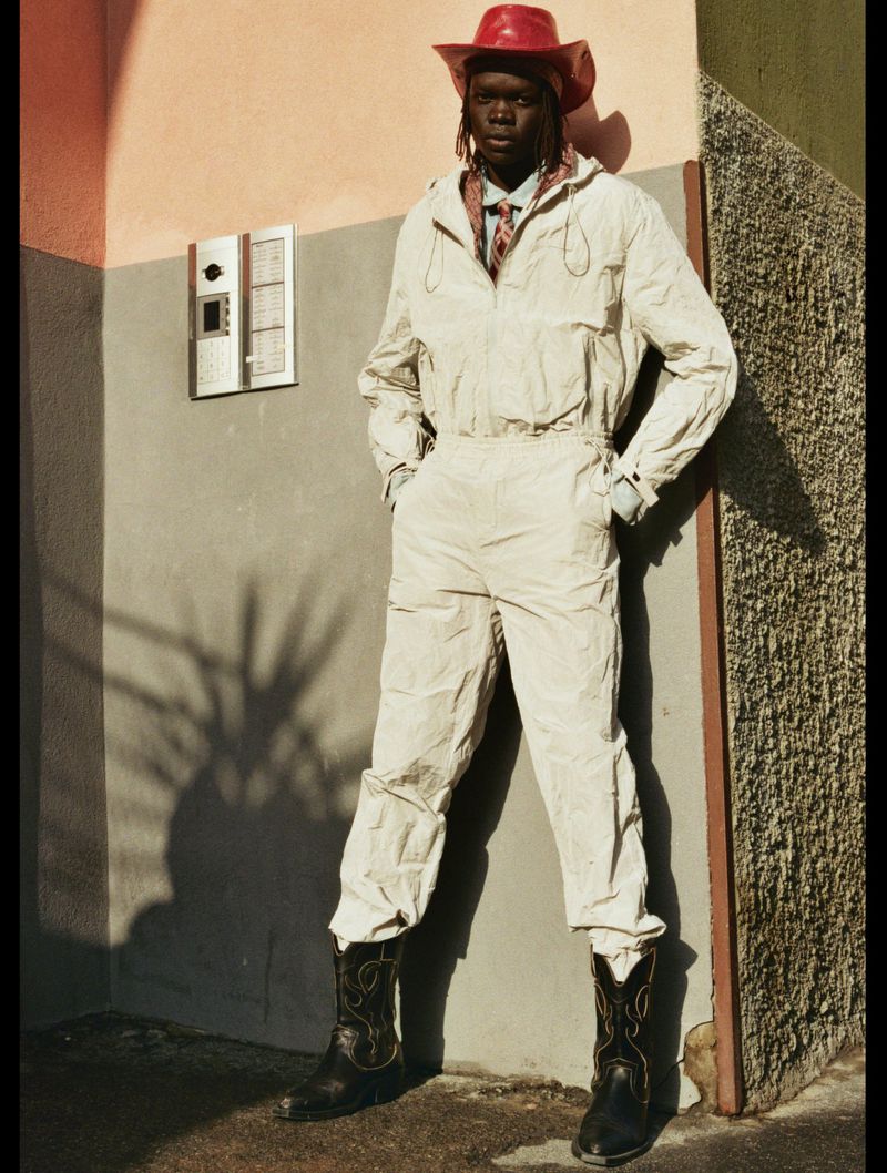 An adult in a light-colored jumpsuit with a red cowboy hat and cowboy boots standing in front of an urban building.