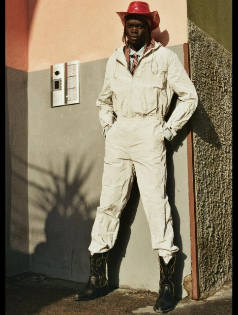 An adult in a light-colored jumpsuit with a red cowboy hat and cowboy boots standing in front of an urban building.