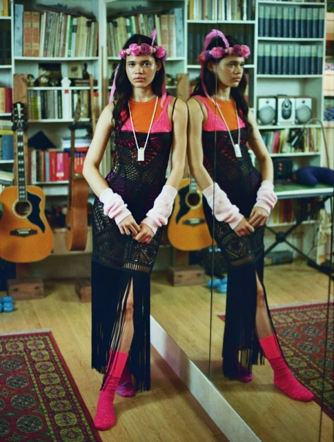Woman in a black knit dress with fringe poses in front of a mirror in a room with books and musical instruments, showcasing a unique fashion style.