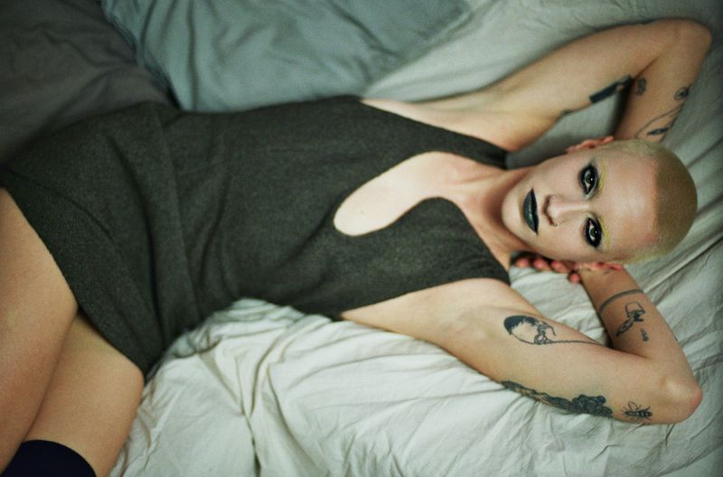 A woman with a shaved head and dark lipstick poses on a bed wearing a short gray dress with a cutout. Her arms are raised above her head.