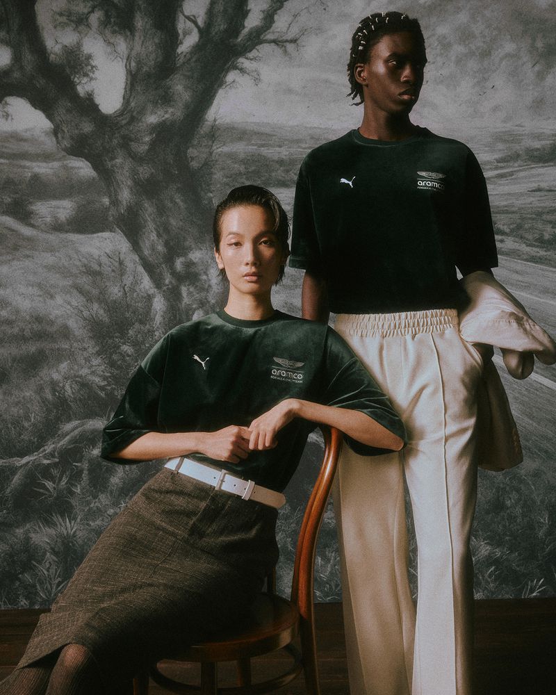 Two models posing in dark green velvet Puma and Aston Martin t-shirts against a scenic painted landscape backdrop.