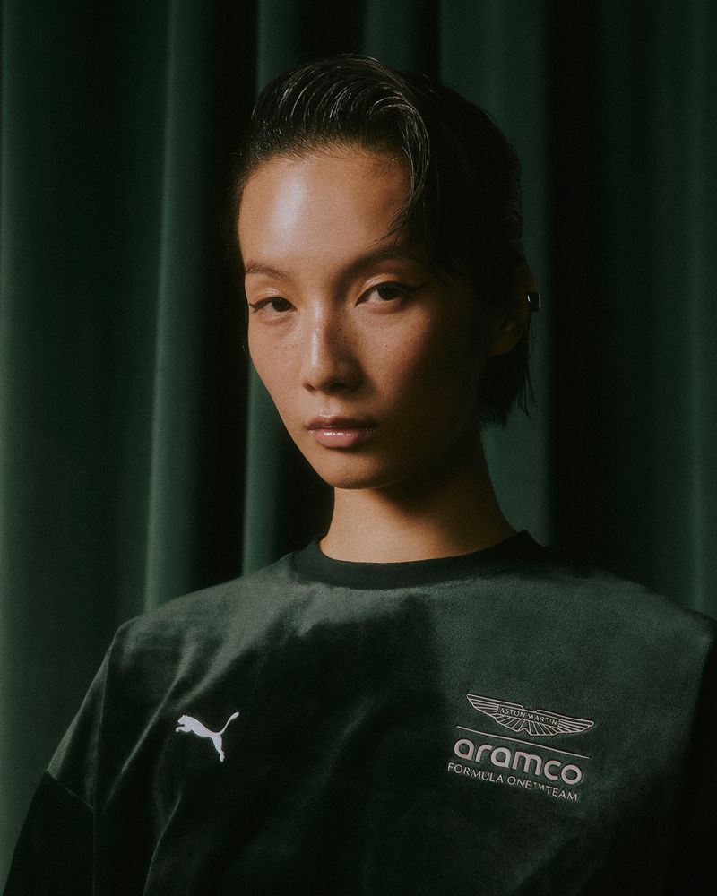 East Asian model wearing a dark green velvet Aston Martin F1 and Puma crewneck sweatshirt against green pleated curtains.
