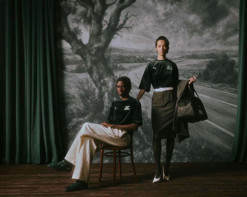 Two models posing in a styled set with a large painted backdrop depicting a tree and road, wearing Puma and Aston Martin F1 apparel.