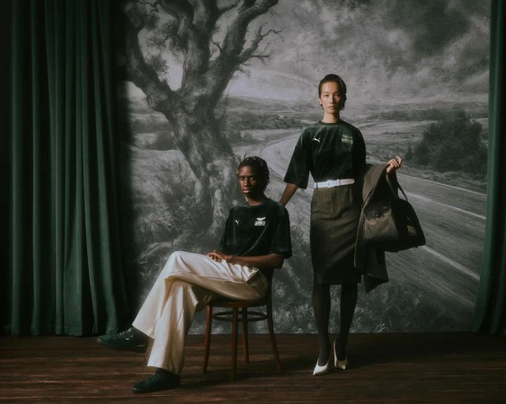 Two models posing in a styled set with a large painted backdrop depicting a tree and road, wearing Puma and Aston Martin F1 apparel.