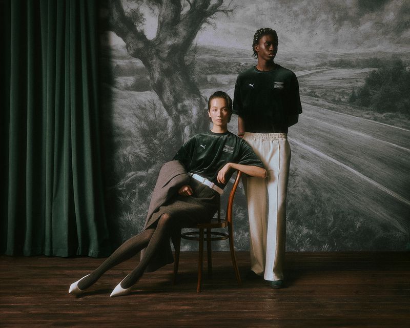 Two models posed against a large painted landscape backdrop, a seated woman and standing man wearing dark shirts and light trousers.