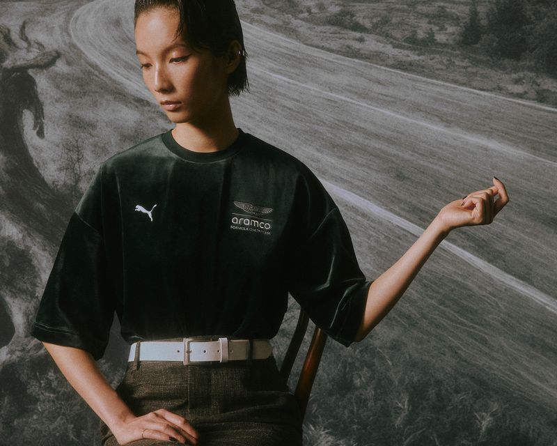 Woman wearing a dark green velvet Puma Aston Martin F1 t-shirt with a white belt against a scenic printed backdrop