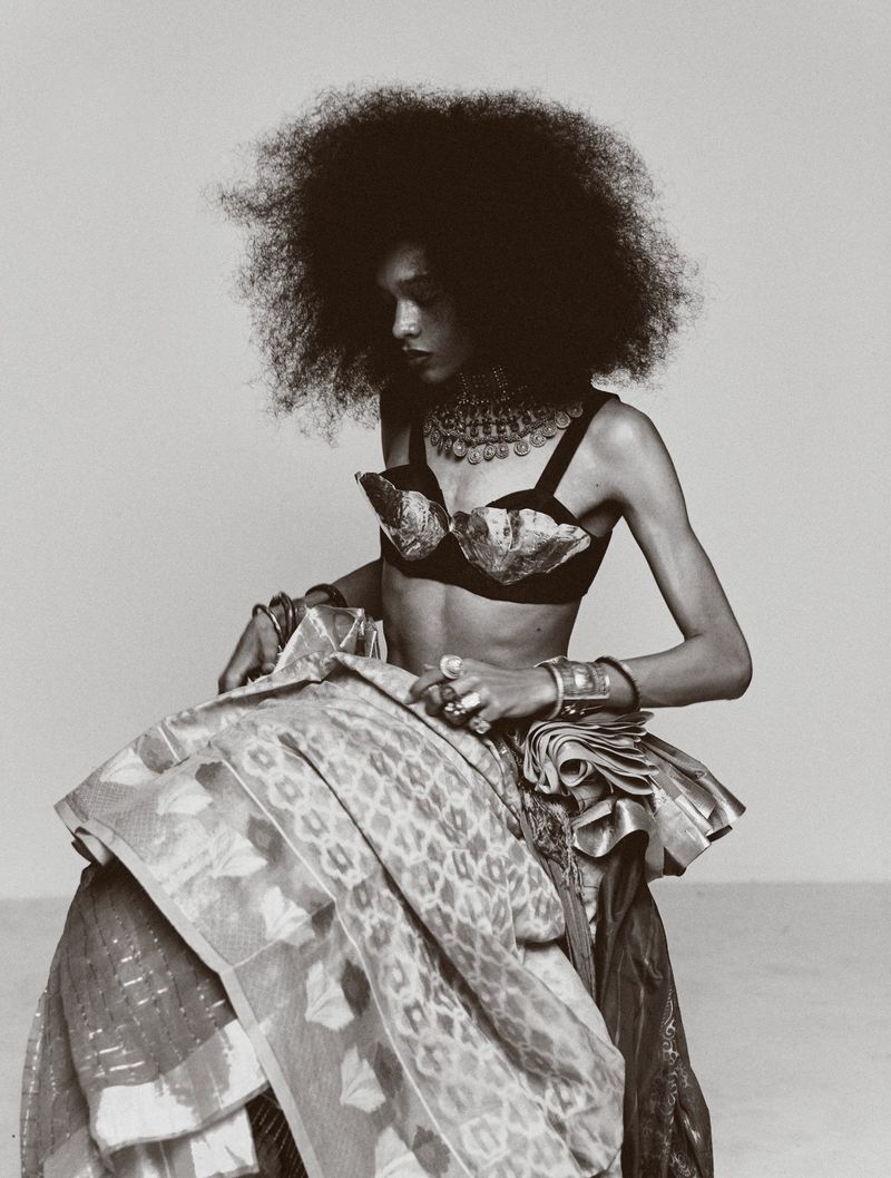 Striking portrait of a young Black woman with large Afro, ornate jewelry, and patterned fabric seated against a neutral backdrop.