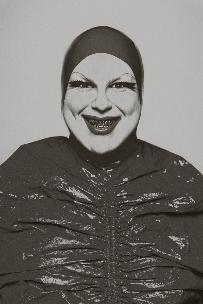 Close-up studio shot of a smiling person with exaggerated makeup, wearing a shiny dark top and matching head covering, against a gray backdrop.