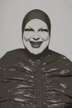 Close-up studio shot of a smiling person with exaggerated makeup, wearing a shiny dark top and matching head covering, against a gray backdrop.