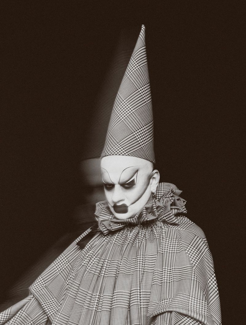 Person in clown costume with white face makeup, dramatic dark eyes, and plaid pointed hat against a dark background in a studio.