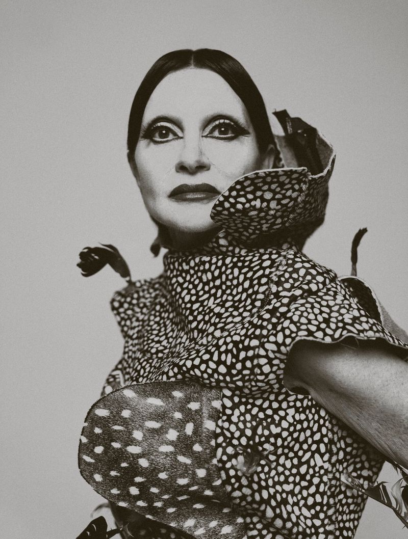 Monochrome portrait of a woman in a spotted dress with dramatic makeup, looking directly at the camera against a plain background.