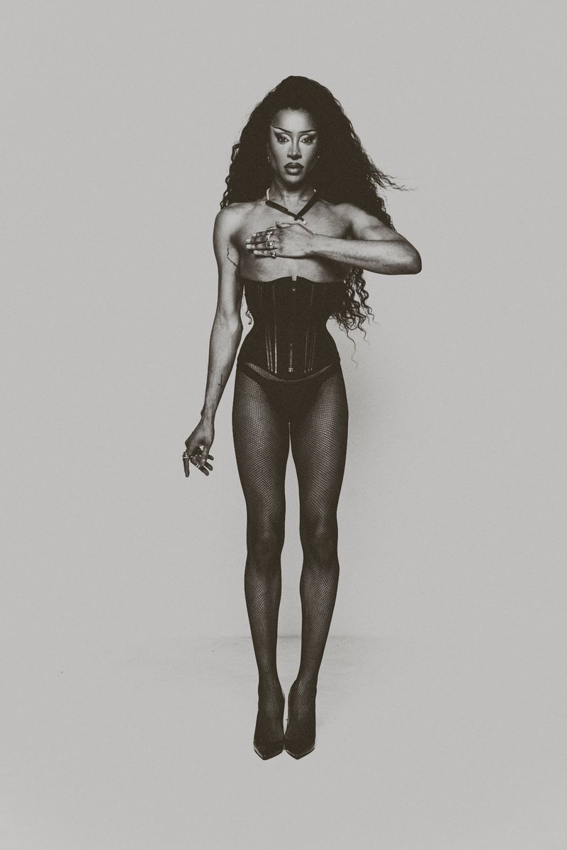 Drag queen posing in a studio wearing a black corset, panty and fishnet stocking with long wavy dark hair and holding her right hand up.