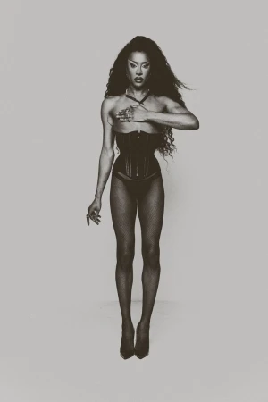 Drag queen posing in a studio wearing a black corset, panty and fishnet stocking with long wavy dark hair and holding her right hand up.