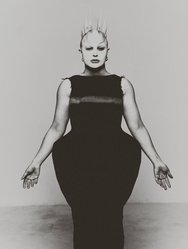 Striking studio portrait of a bald adult female with spiky hair extensions, wearing a unique black dress and jewelry, set against a gray background.