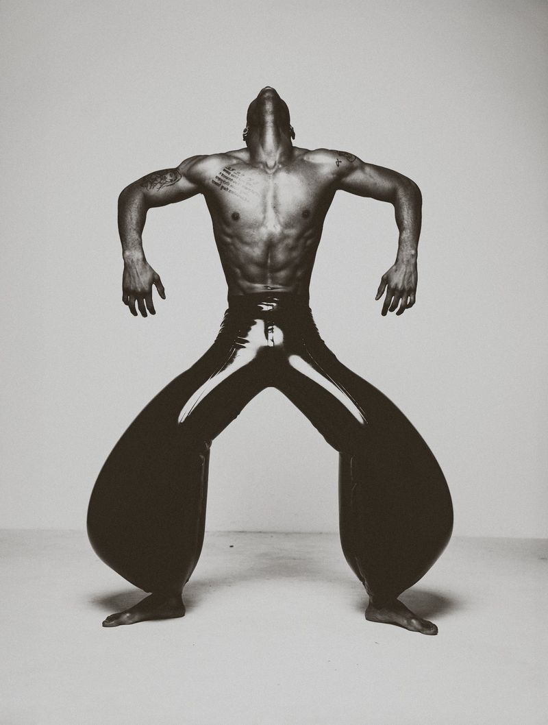 Athletic build African-American man with bare chest and shiny wide-leg pants in a dynamic pose against a neutral backdrop.