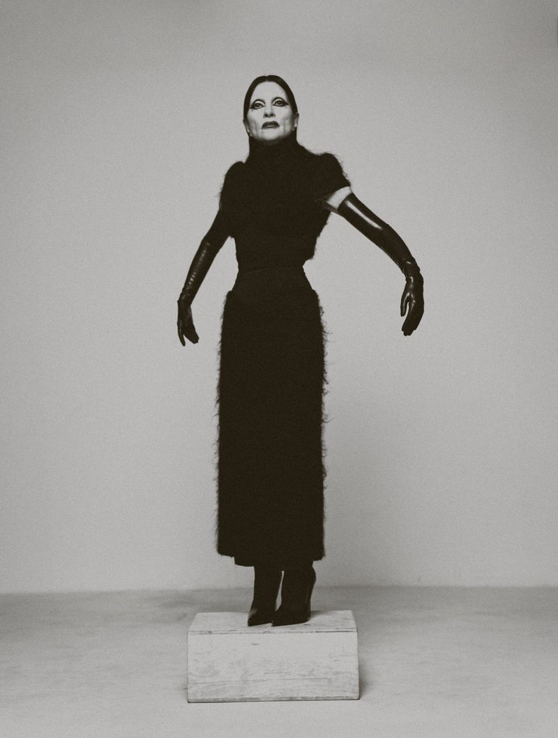 Monochrome portrait of an adult female standing on a platform in a long black dress and elbow-length gloves against a plain background.