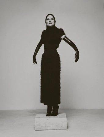 Monochrome portrait of an adult female standing on a platform in a long black dress and elbow-length gloves against a plain background.