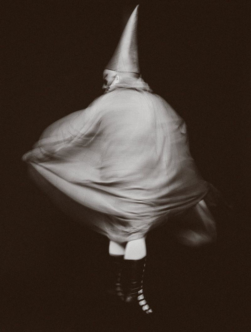 Person in pointed hat and flowing cape, action shot with motion blur, dark background, eye-level view, monochrome effect, fashion photography.