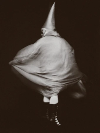 Person in pointed hat and flowing cape, action shot with motion blur, dark background, eye-level view, monochrome effect, fashion photography.